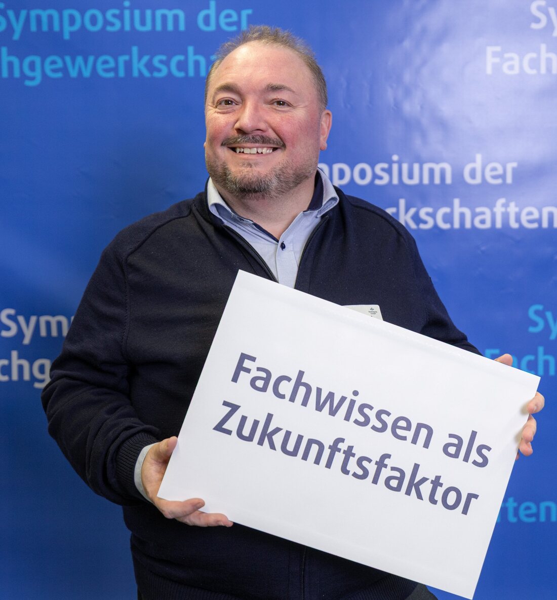 Benjamin Swierkot hält ein Schild mit der Aufschrift „Fachwissen als Zukunftsfaktor“ beim Symposium der Fachgewerkschaften
